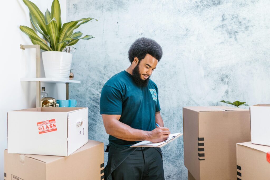 Un hombre con barba y camiseta de mudanzas revisa una lista de inventario en un portapapeles, rodeado de cajas de cartón