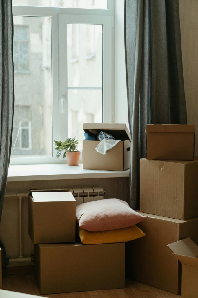 Cajas de mudanza apiladas junto a una ventana con cortinas grises, un radiador y una pequeña planta, simbolizando un espacio recién habitado o en proceso de mudanza
