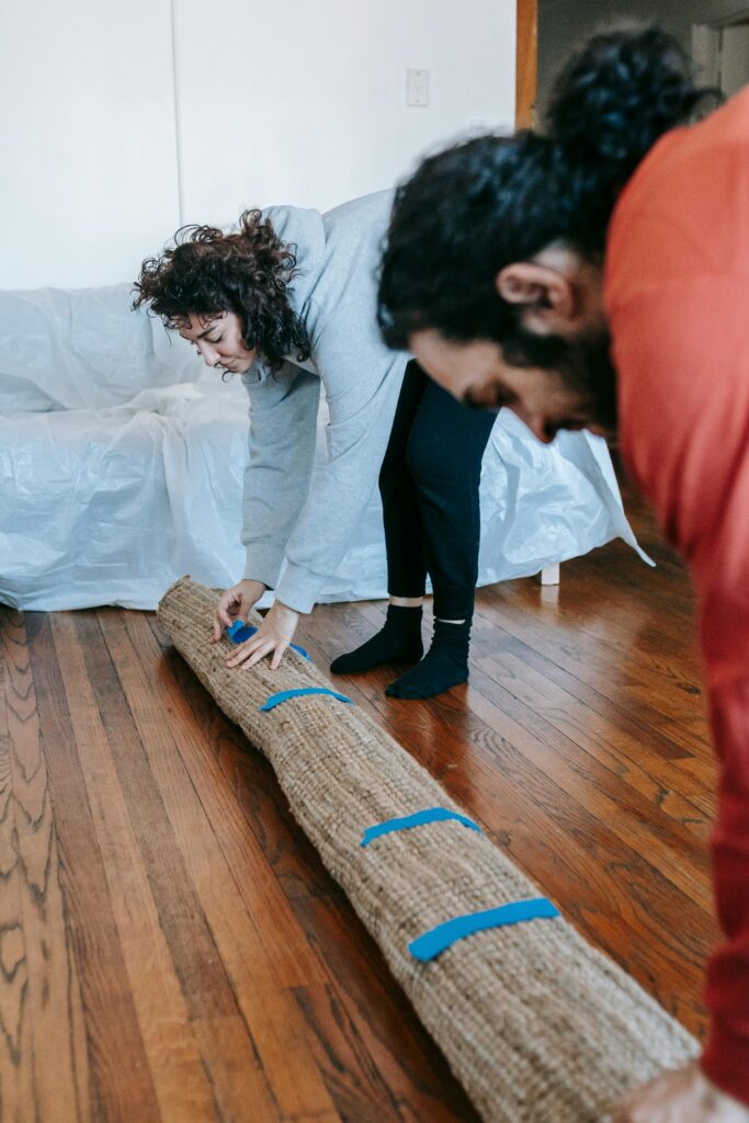 Pareja enrollando alfombra de yute con cinta azul en dormitorio con suelo de madera para almacenamiento